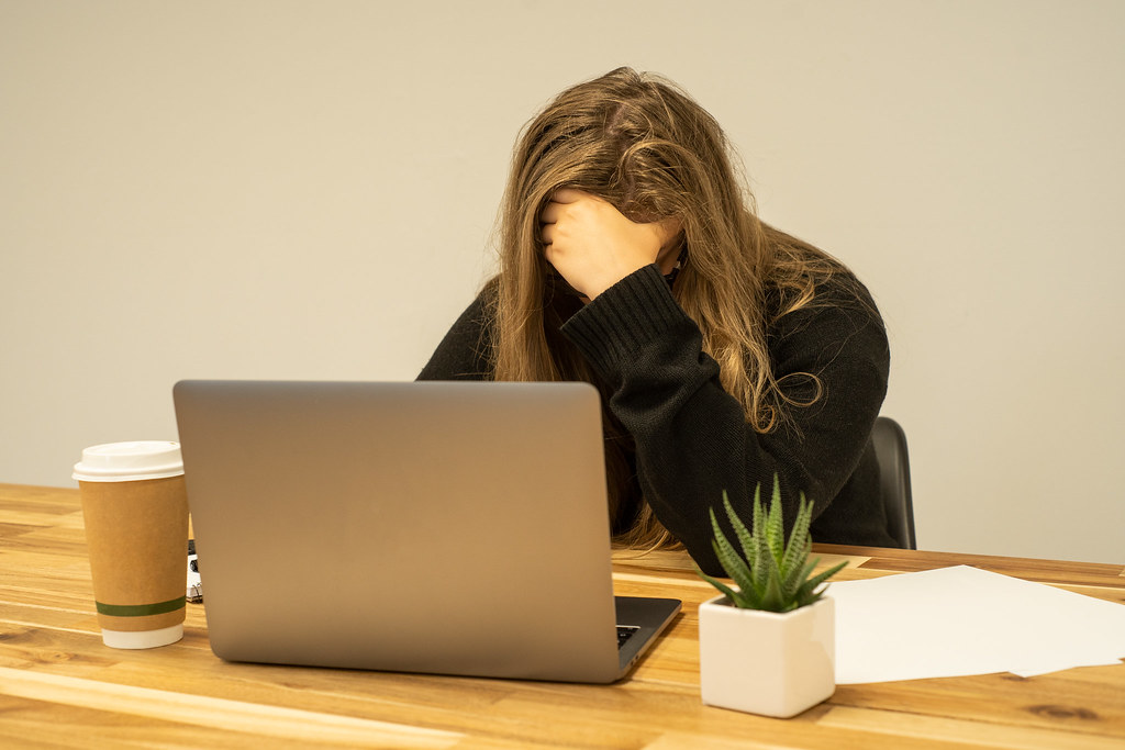 Woman sitting in front of her laptop holding her head, showing clear signs of burnout at work such as stress, mental exhaustion, and overwhelm.